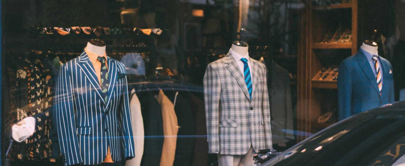 An image of suits through a shopping window - follow this guide to learn everything you need to know about suit trousers.